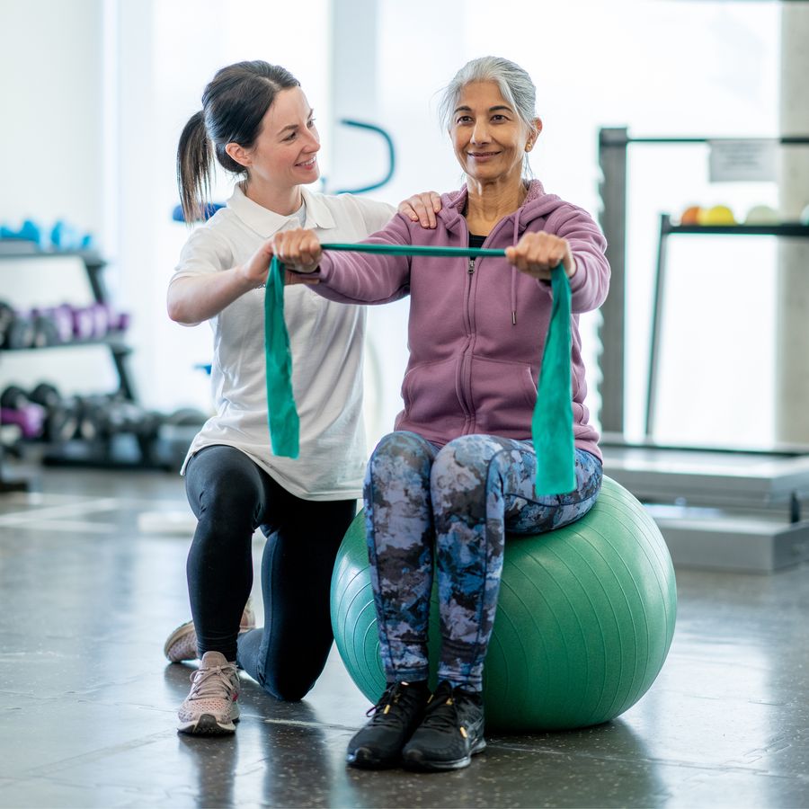 Physiotherapeutin begleitet eine Patientin bei einer Übung mit dem Theraband auf dem Gymnastikball.